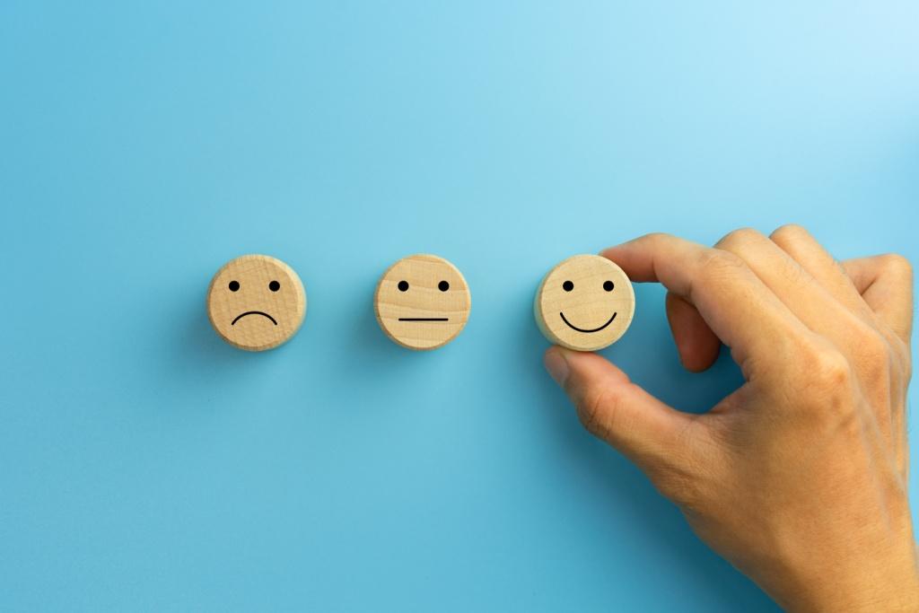 Drei Smileys aus Holz mit jeweils einem traurigen, neutralen und fröhlichen Ausdruck liegen auf einem hellblauen Hintergrund.