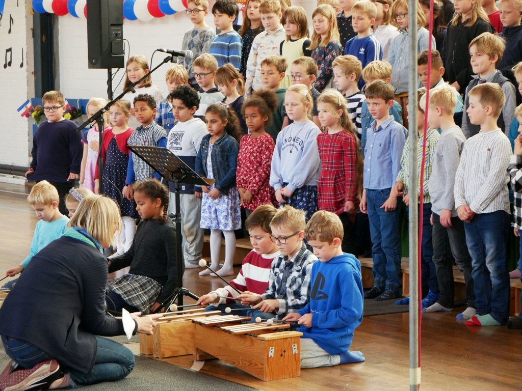 Kinder spielen Xylophon und singen, vor ihnen kniet eine Lehrkraft